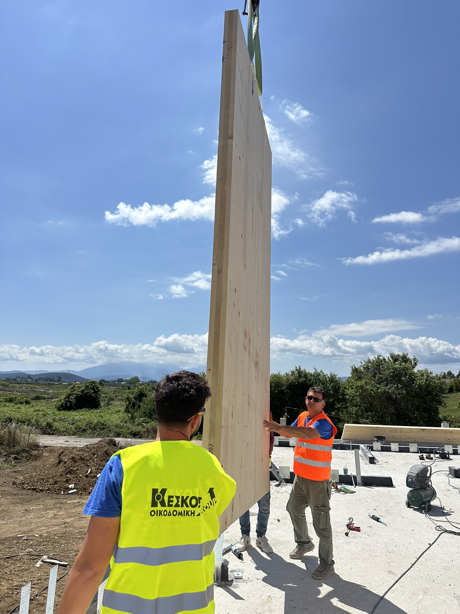 Crane lifts vertical CLT wall at construction site, blue sky, Ionian